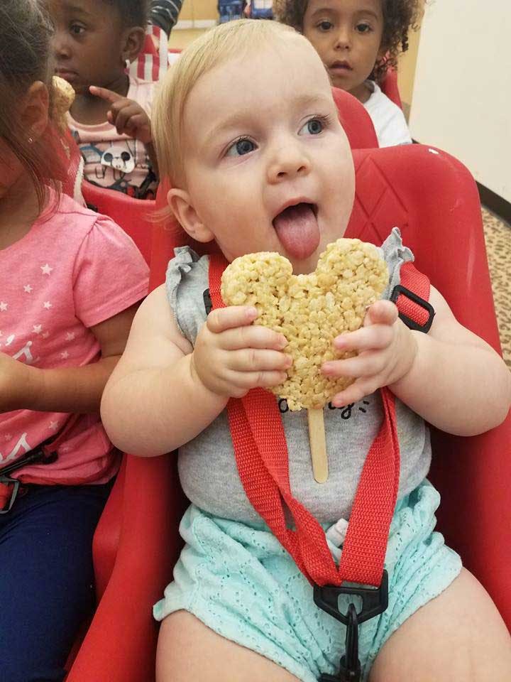Child in a high chair holding a rice crispy treat, tongue out