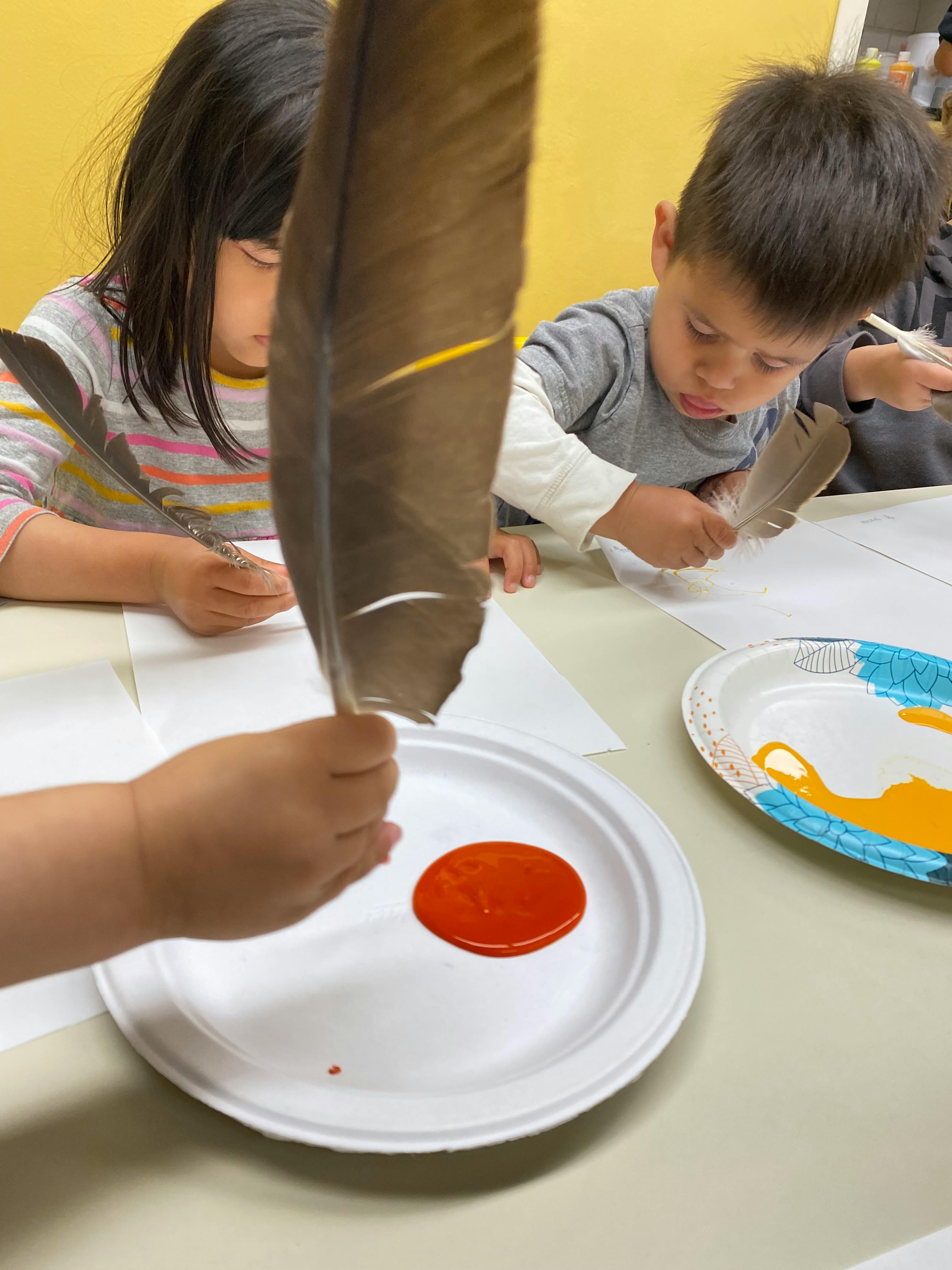 Children using feathers to paint on paper at a table