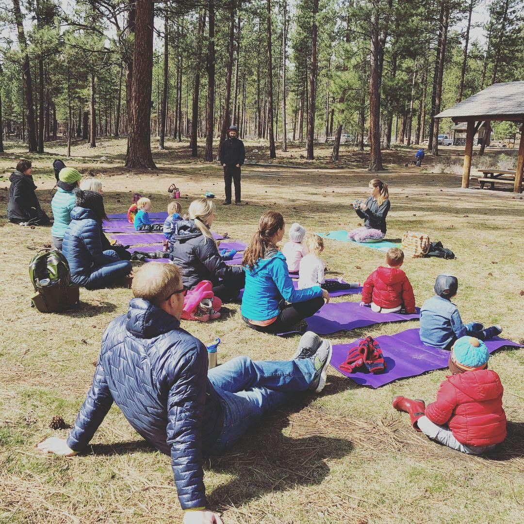 People and children sitting on mats outdoors in a wooded area