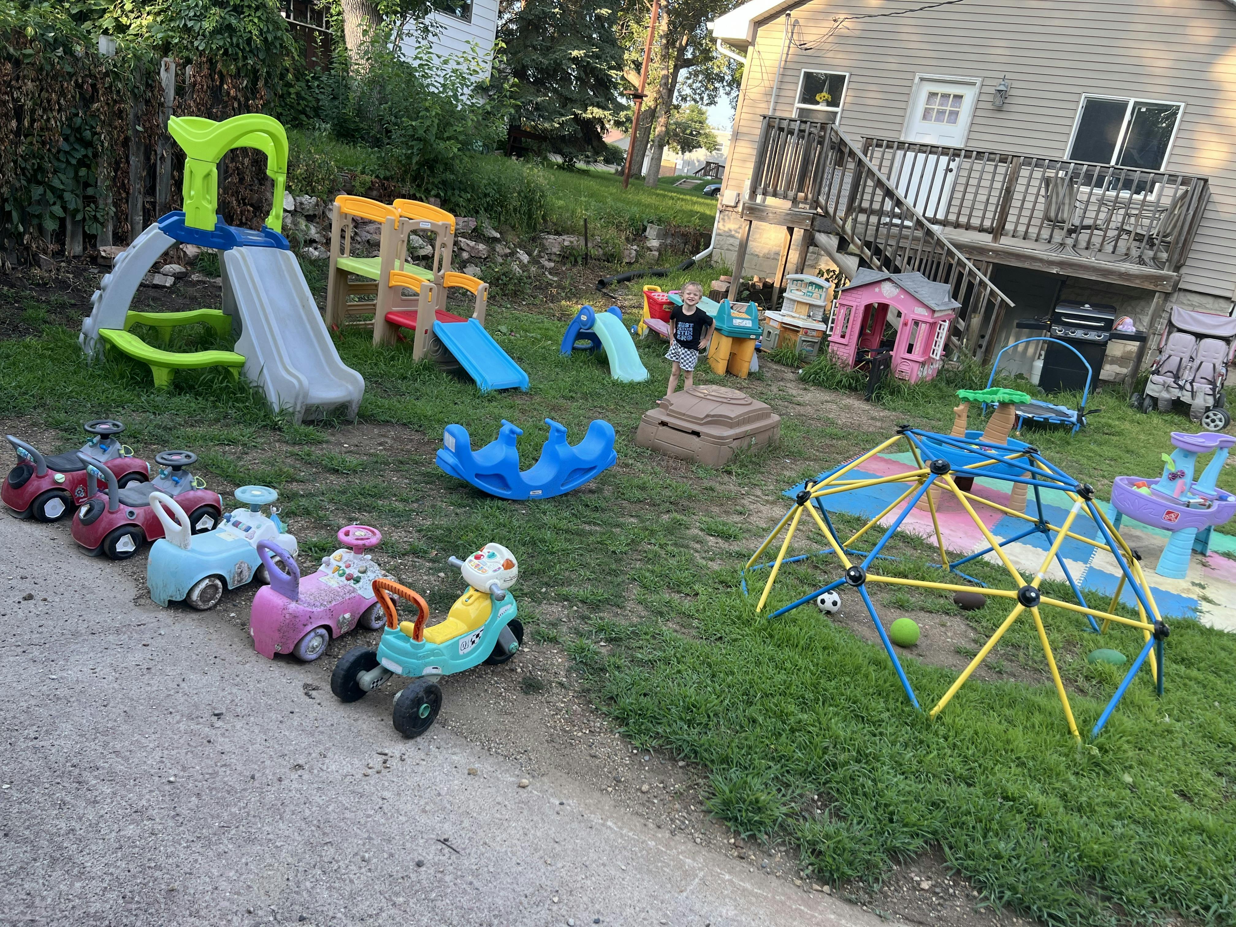 Child standing in a backyard full of colorful play equipment and ride-on toys
