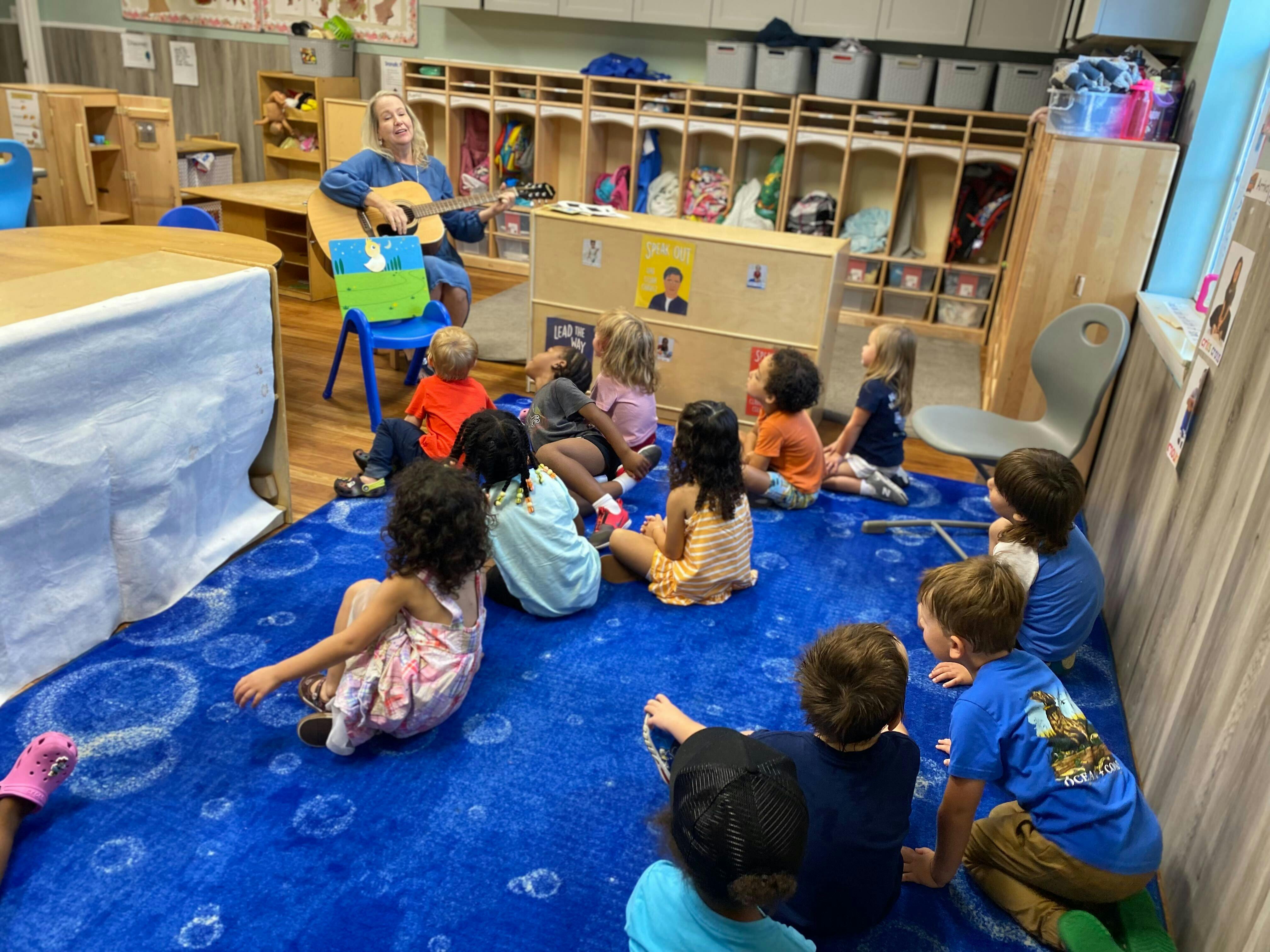 Person playing guitar for a group of children sitting on a blue rug