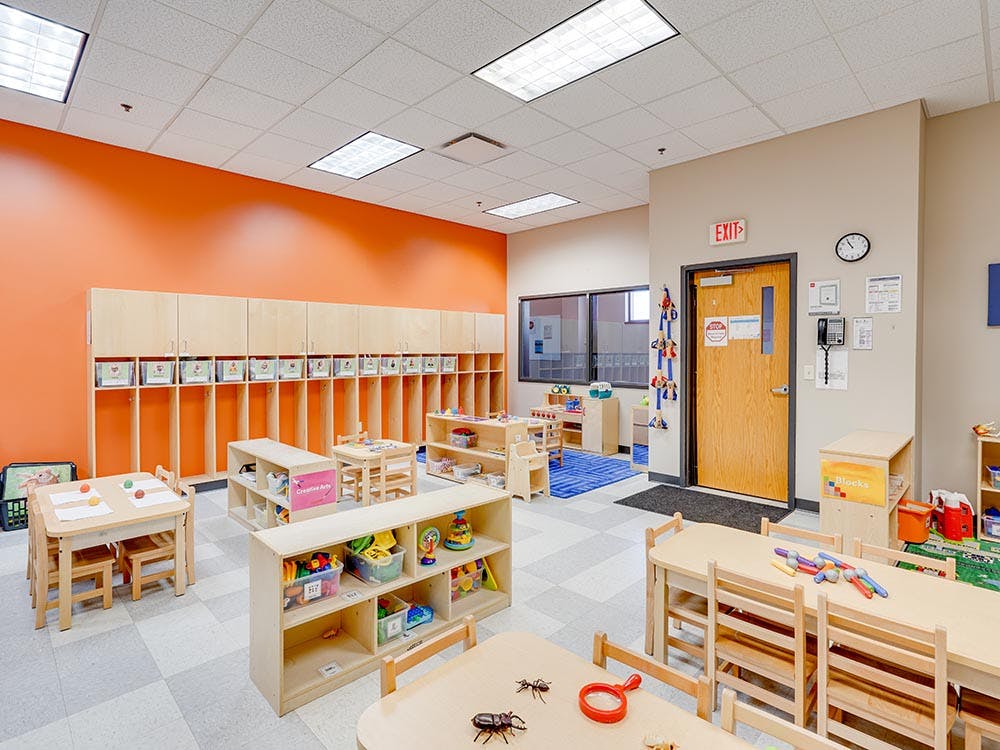 Empty classroom with small tables and chairs, cubbies, and an orange accent wall