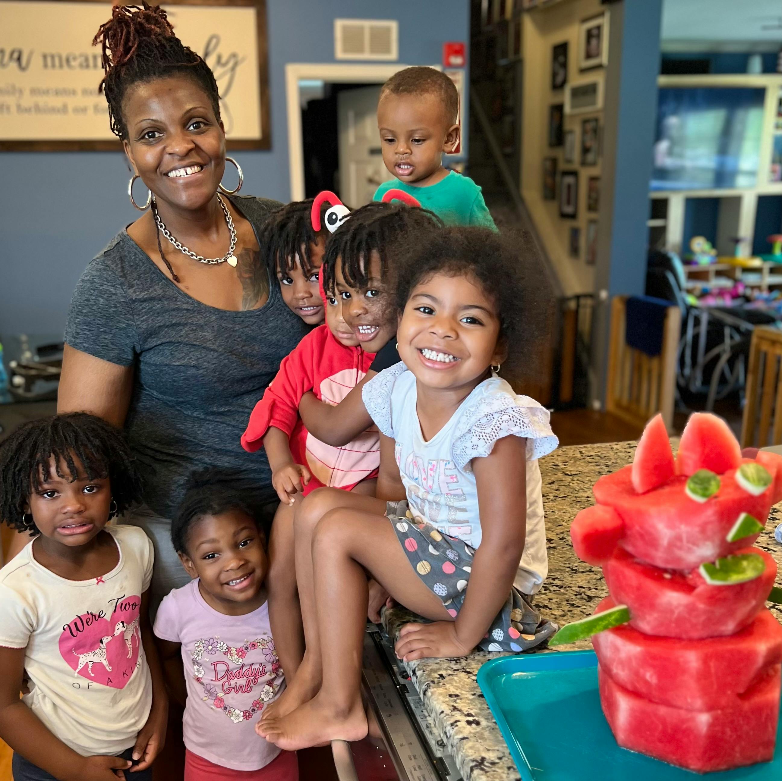 Person with five children, one child sitting on a counter next to a watermelon sculpture