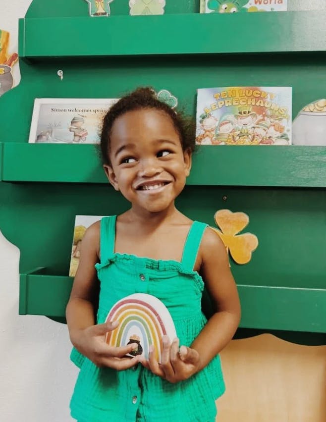Child in green holding a rainbow decoration with a pot of gold