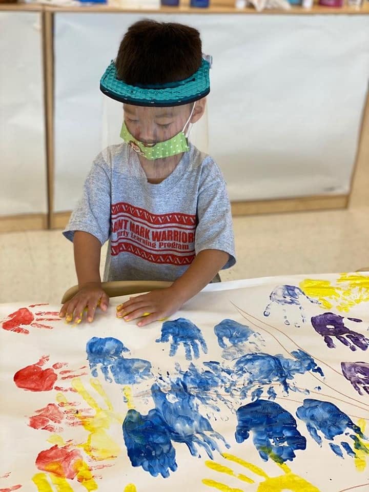 Child in a face shield and mask making handprints on paper