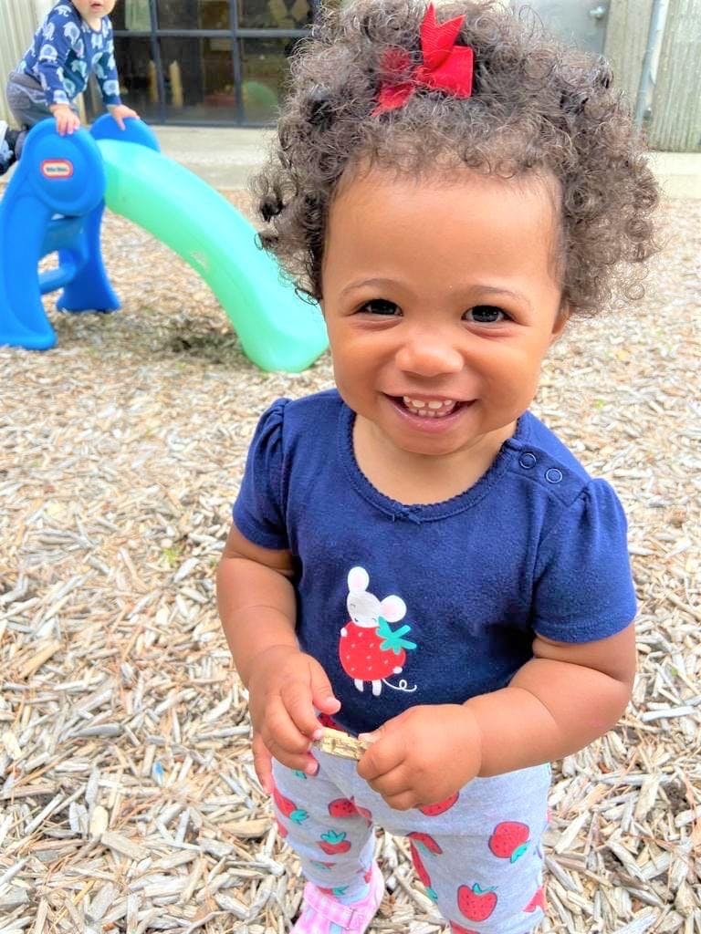 Child with curly hair and red bow smiling at camera, another child on a slide in background