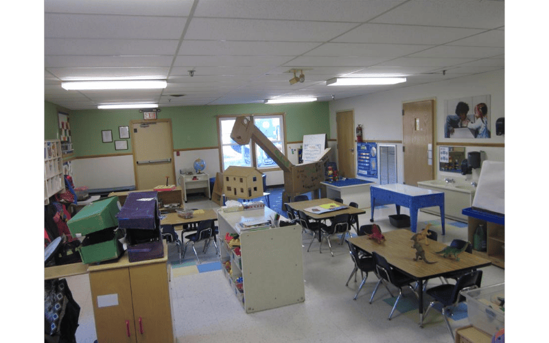 An empty classroom with small tables and chairs, and a large cardboard dinosaur