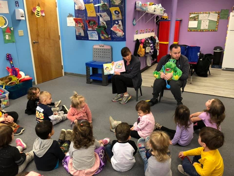 Two adults reading to a group of children sitting on the floor in a classroom