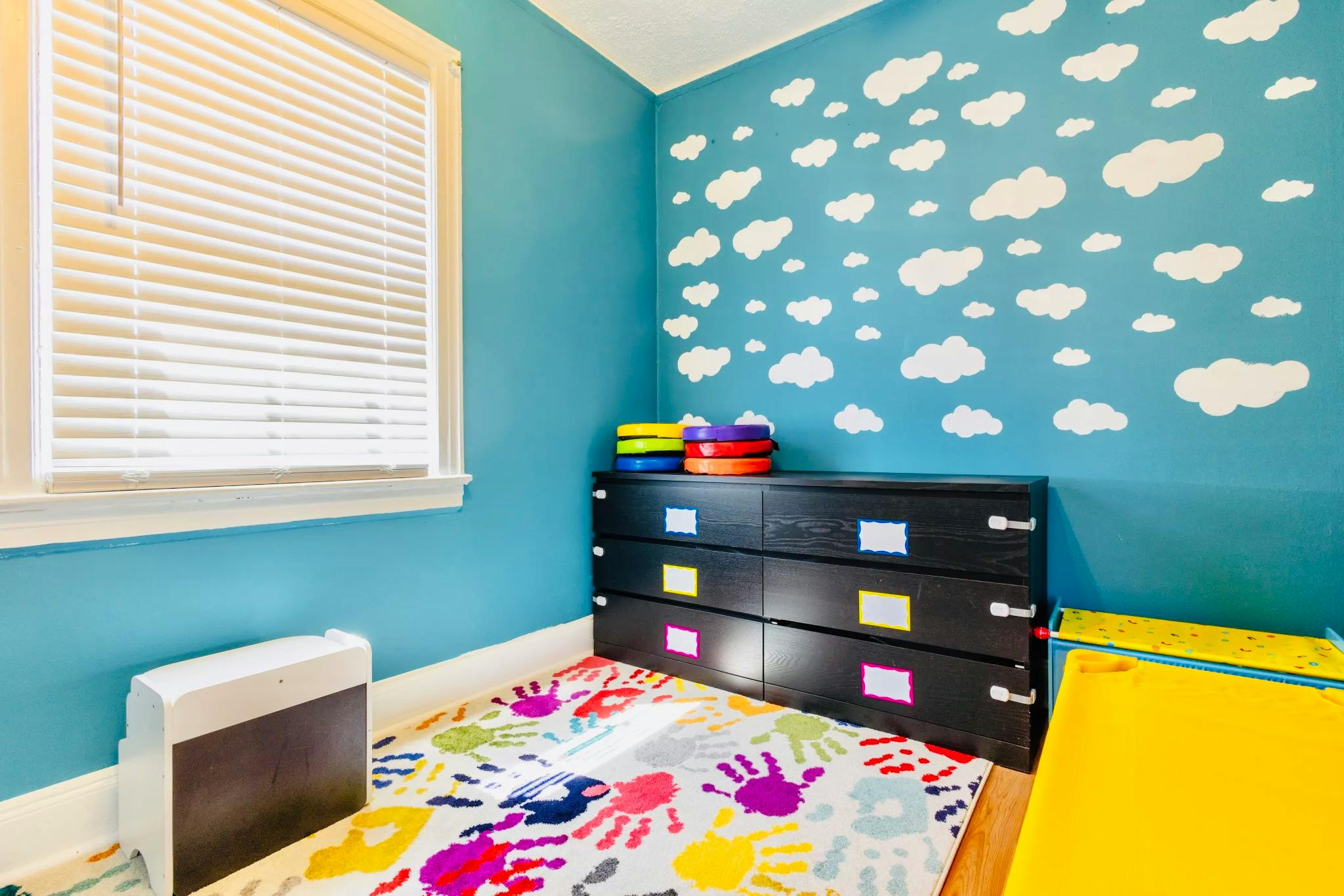Child's room with blue walls, white cloud decals, and a colorful handprint rug