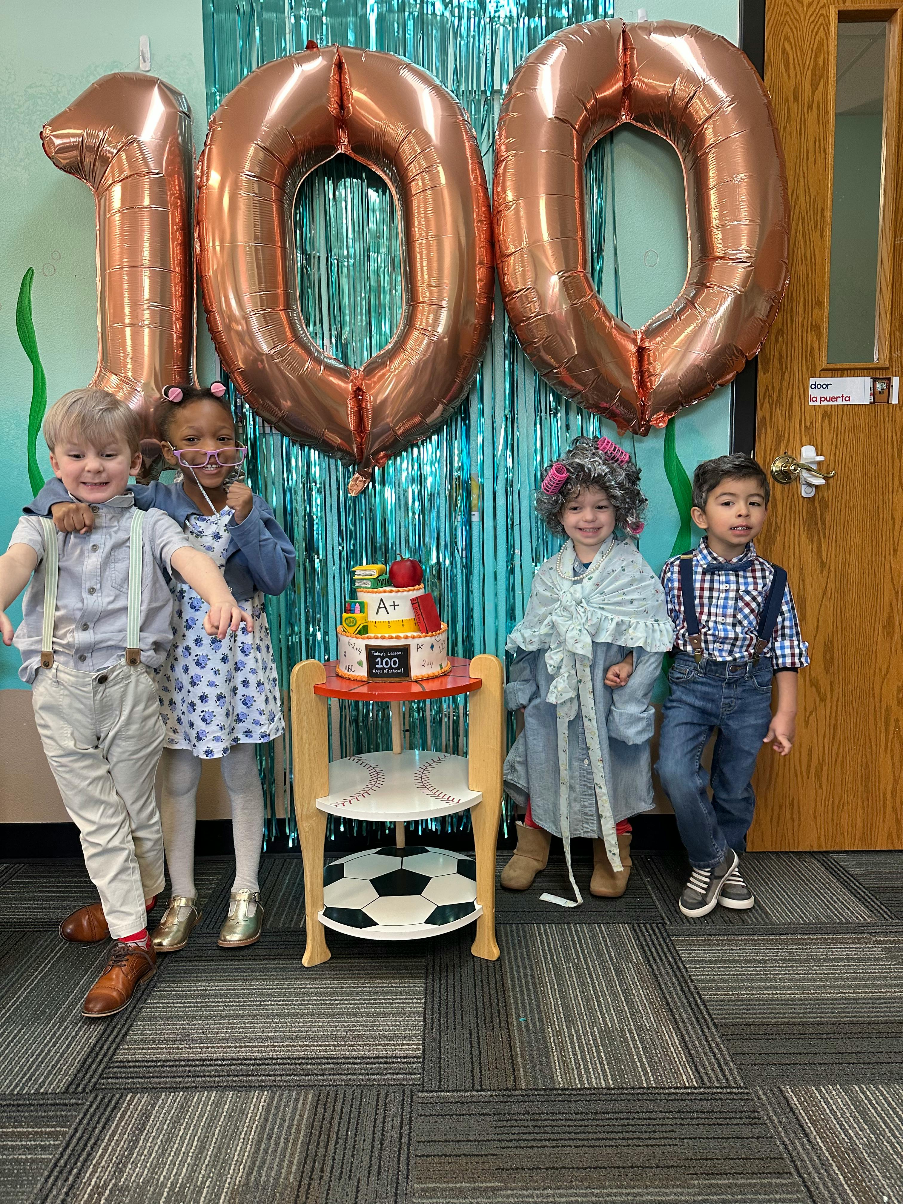Four children standing in front of large 100 balloons and a cake