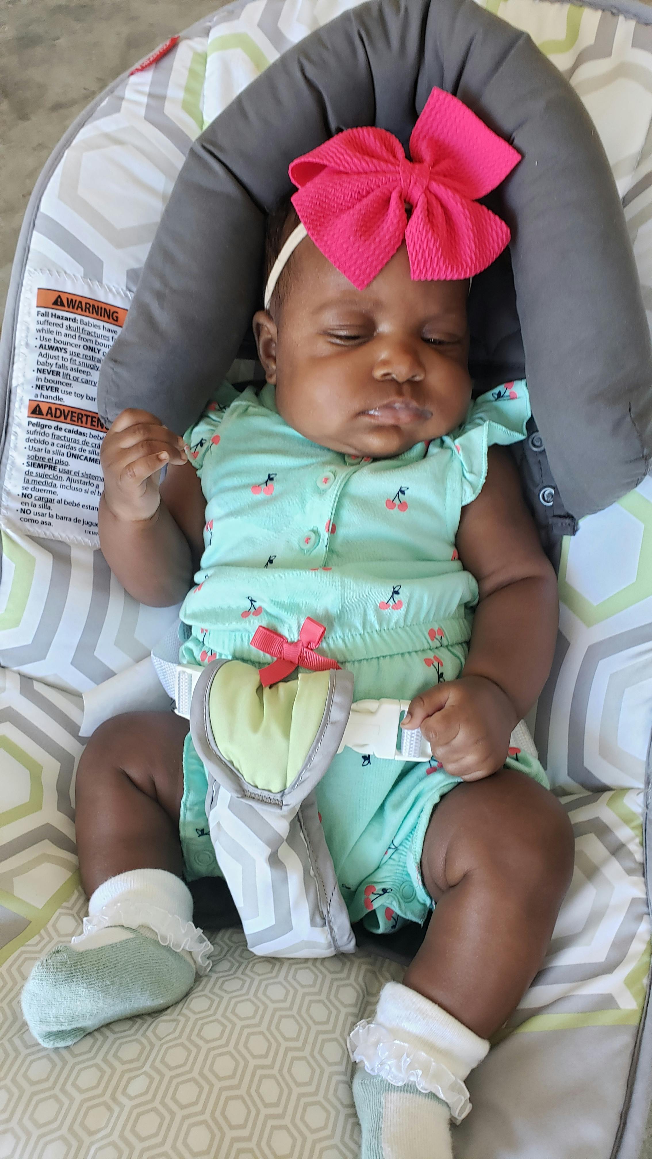 Child sleeping in a baby bouncer with a pink bow on their head