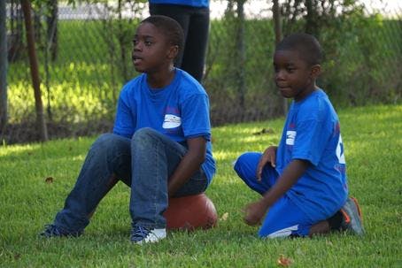 Two children in blue shirts sitting on grass, one on a ball