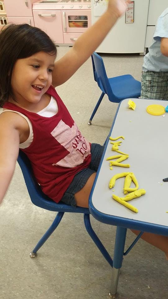 Child sitting at a table with yellow play-doh, raising an arm