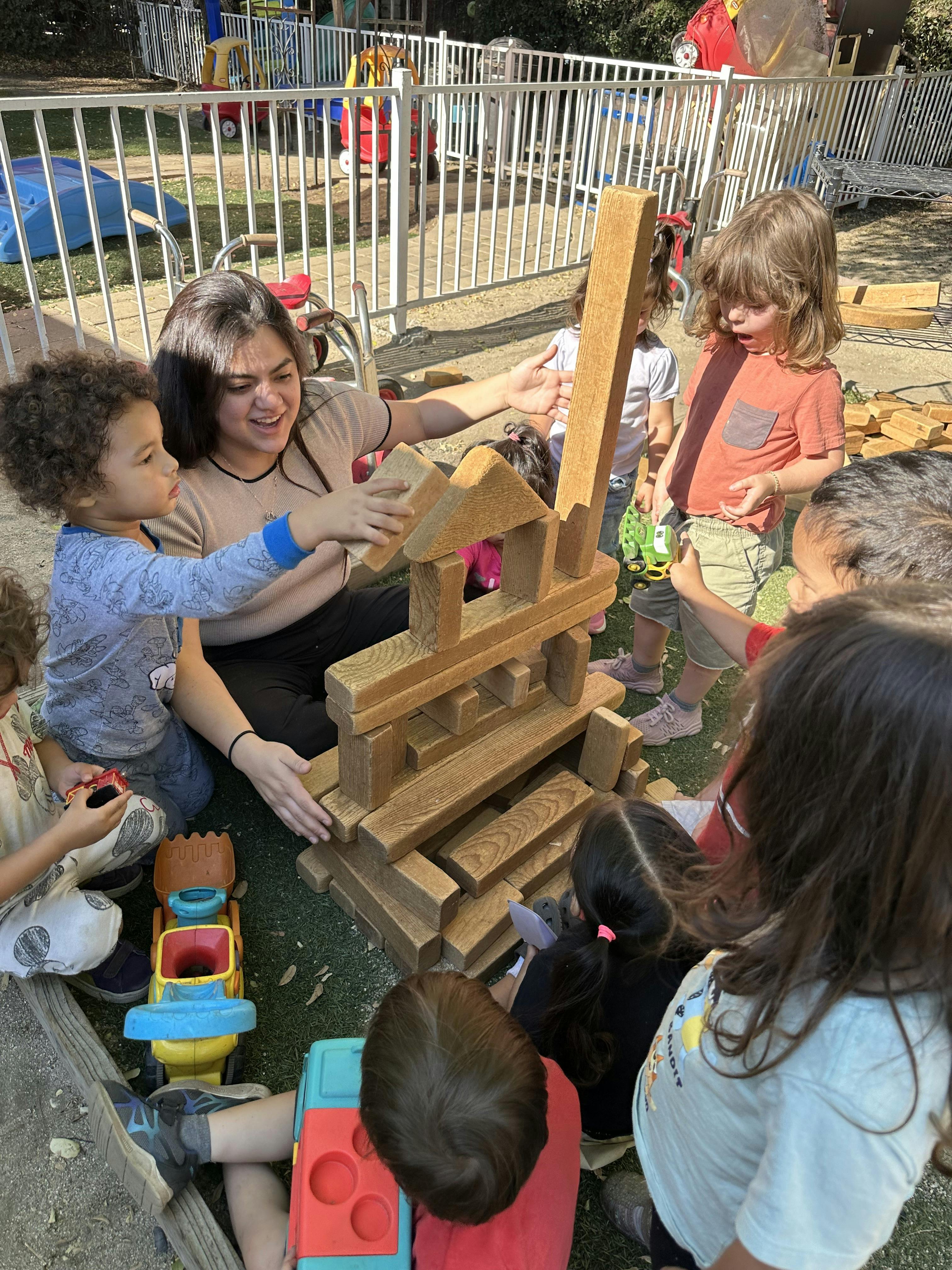 A person and multiple children playing with large wooden blocks on grass