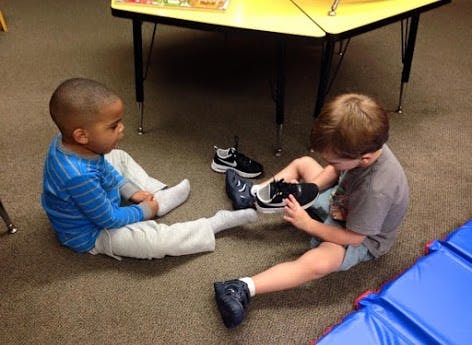 Two children sitting on the floor, one putting on a shoe