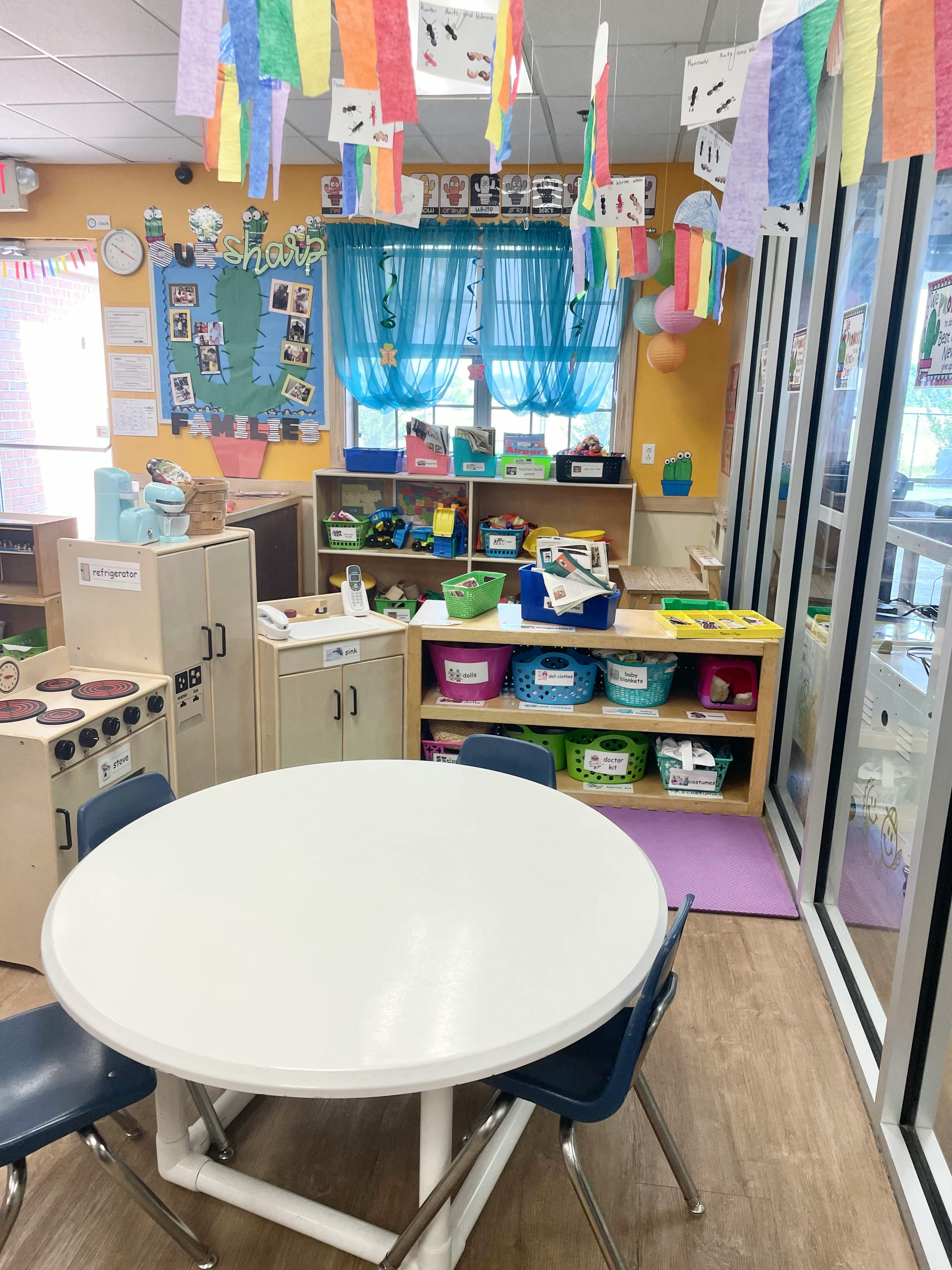 Classroom with a white round table and chairs, shelves with bins, and a play kitchen
