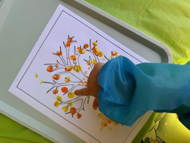 Child's hand in blue sleeve finger painting a tree with orange and yellow dots
