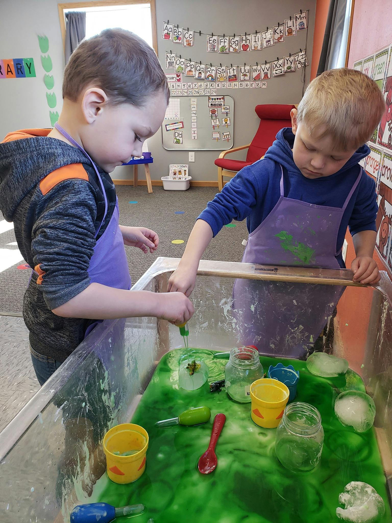 Two children in aprons playing with green slime and toys in a sensory bin
