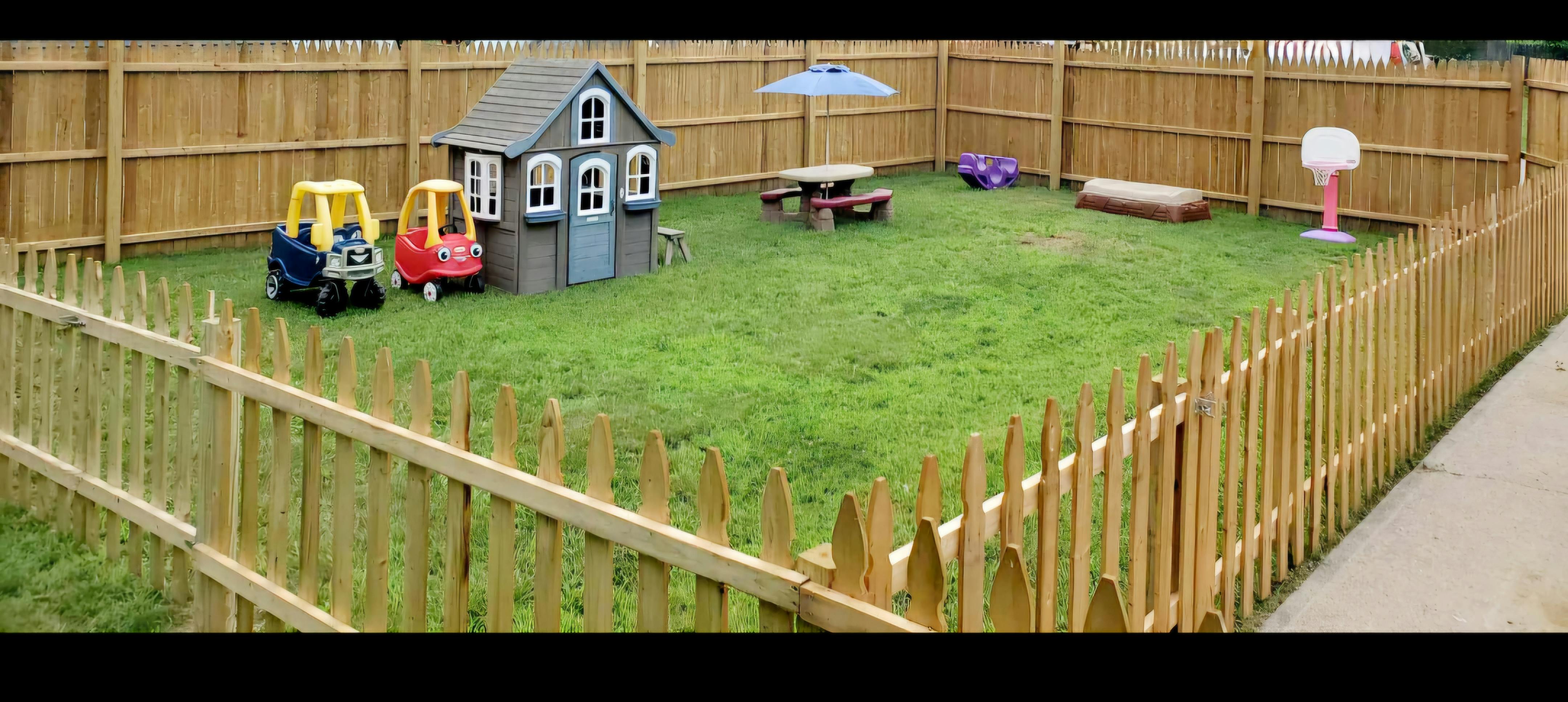 Fenced backyard with playhouse, toy cars, picnic table, and basketball hoop