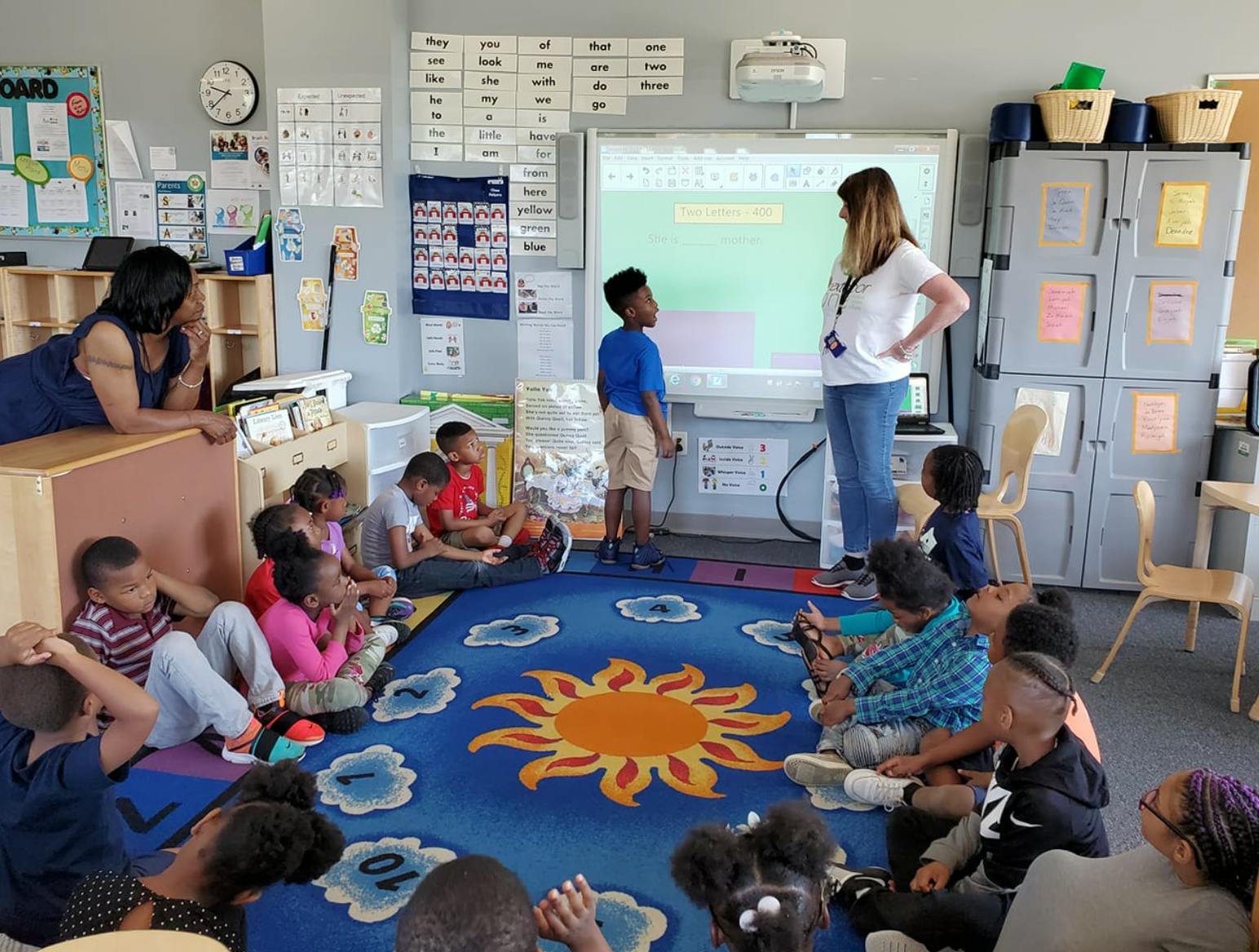 Adults and children in a classroom, with one adult and child at a whiteboard