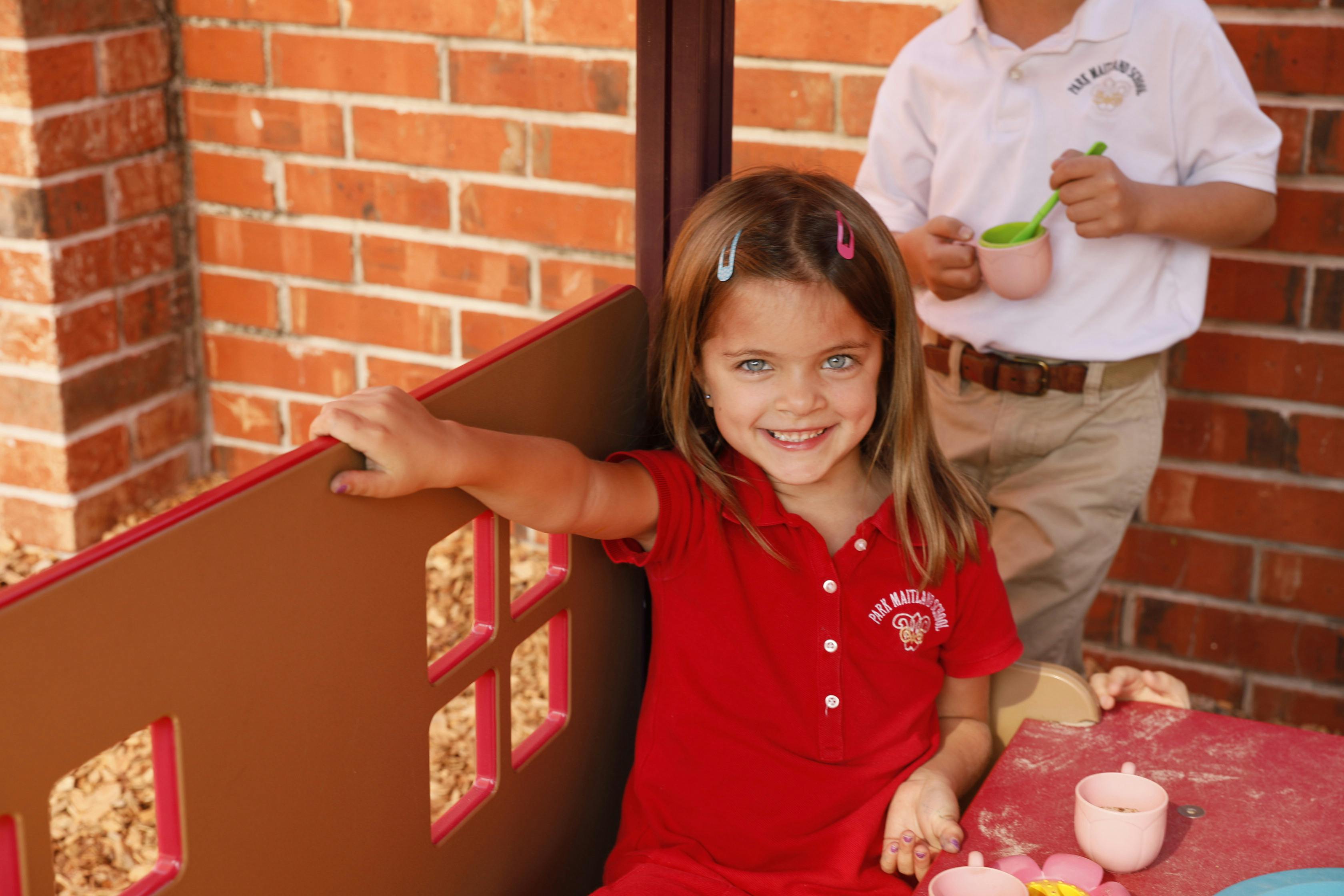 Child in red shirt sitting at a play table, person standing in background