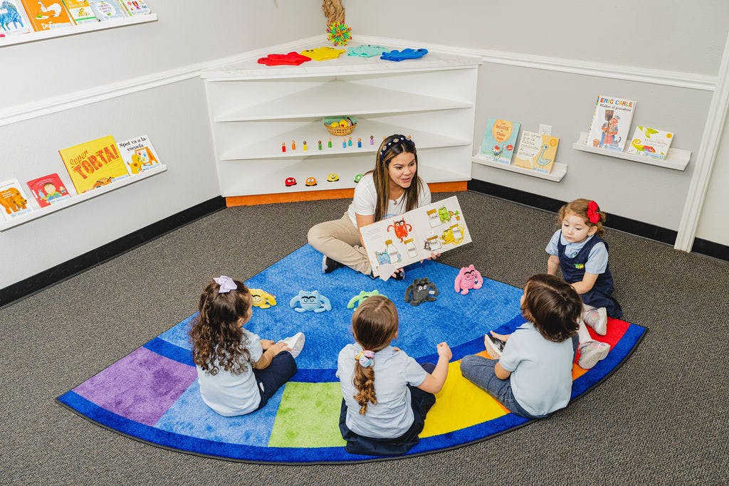 Person reading a book to three children on a colorful rug in a classroom