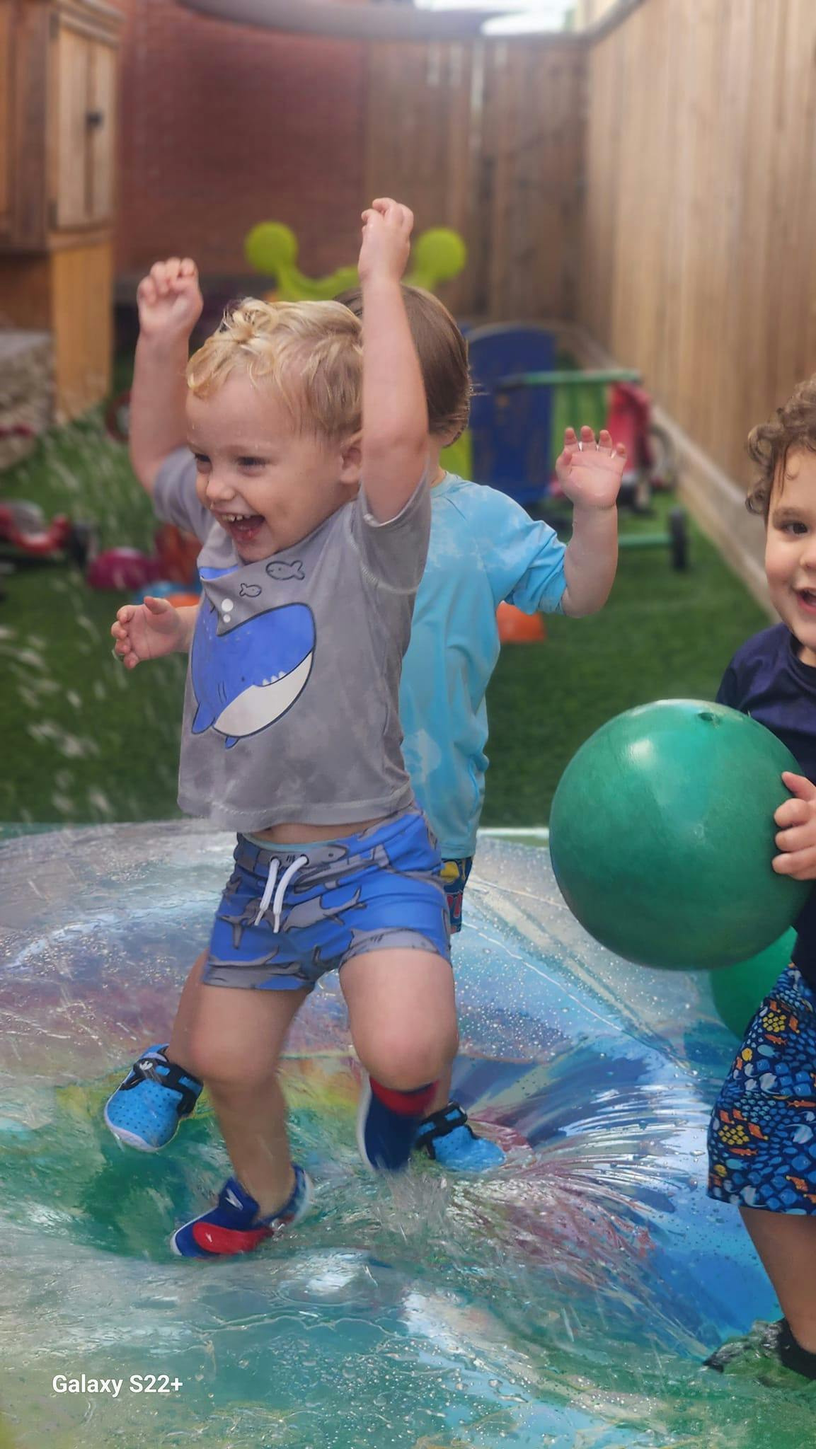 Child with arms raised standing in water, another child holding a green ball