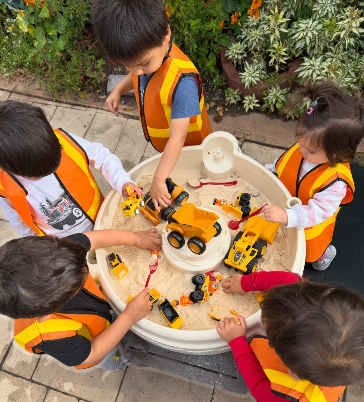 Four children in orange vests playing in a sandpit with toy construction vehicles