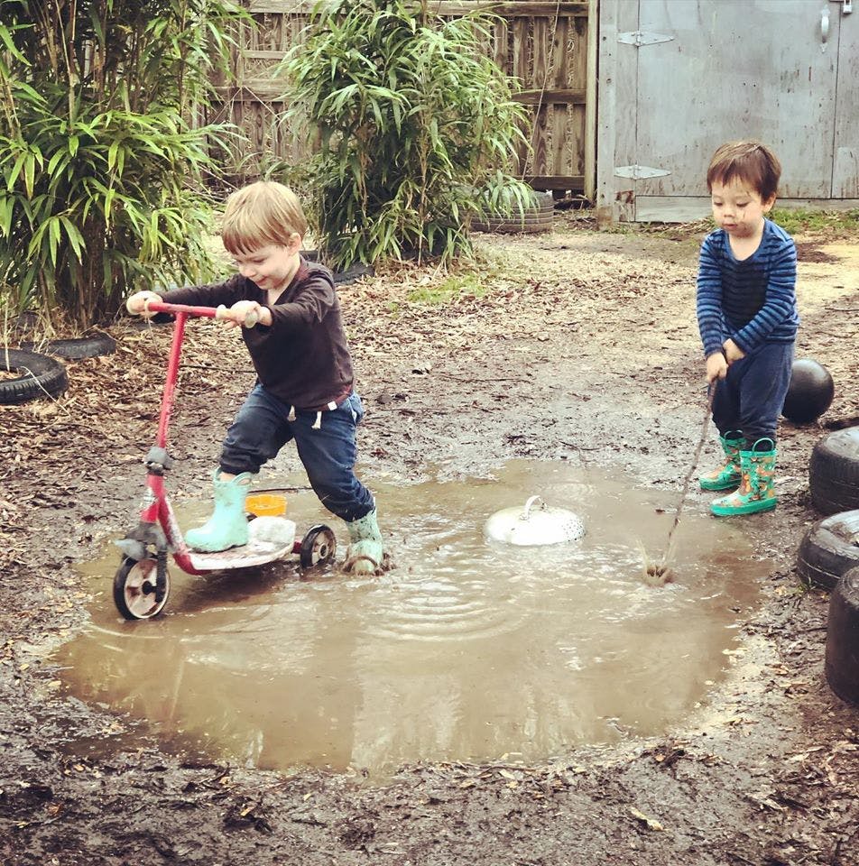 Two children playing in a muddy puddle, one on a scooter and one with a stick