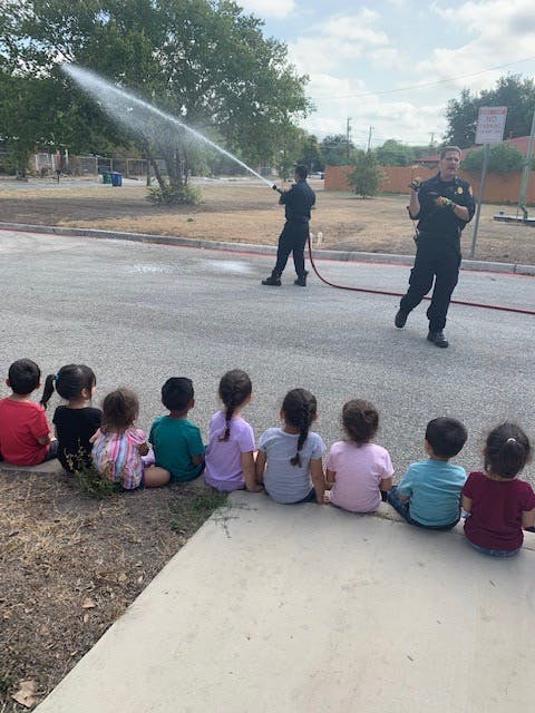 Children sitting on a curb watching a person spray water from a hose