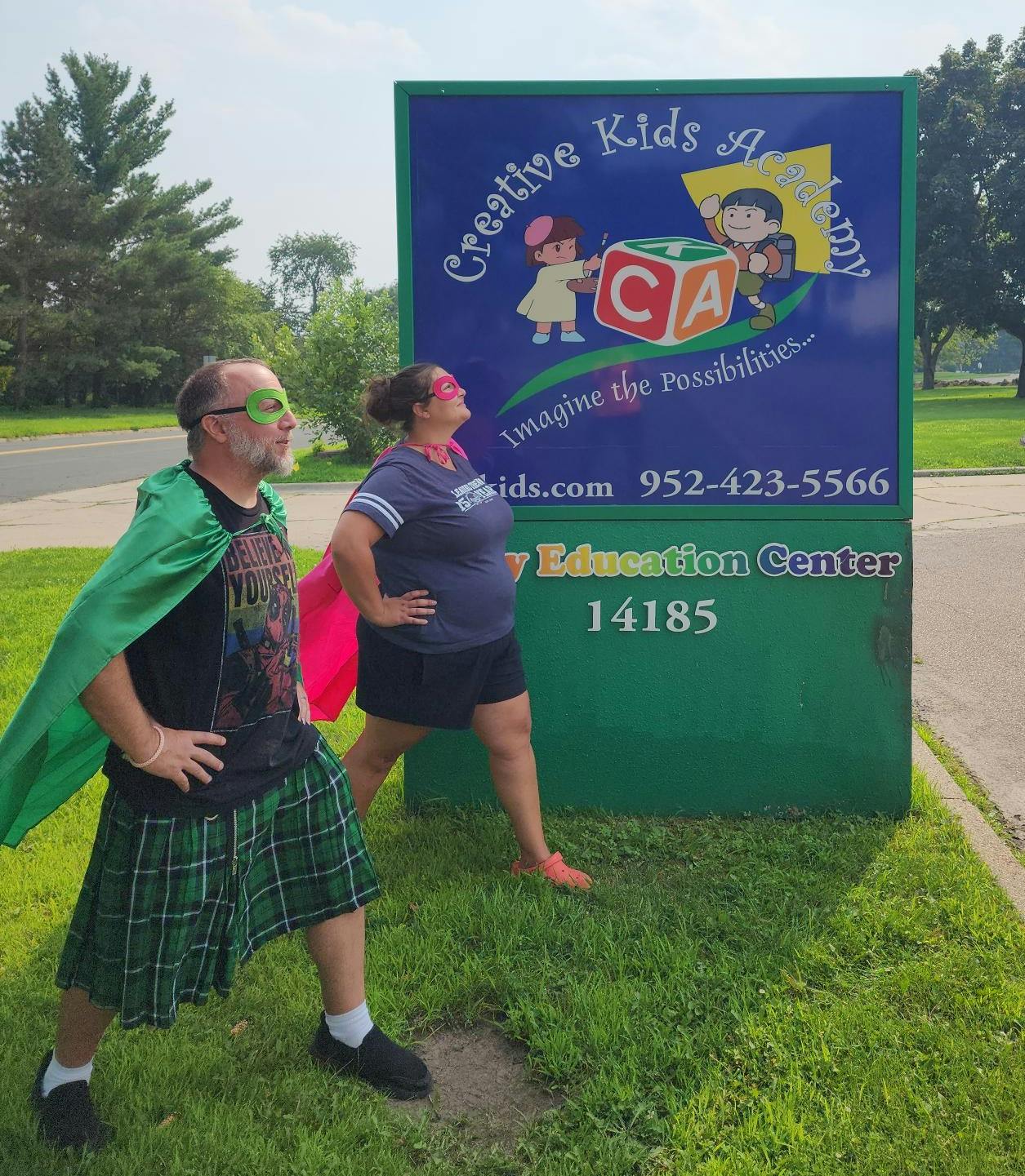 Two people in superhero capes and masks posing in front of an academy sign