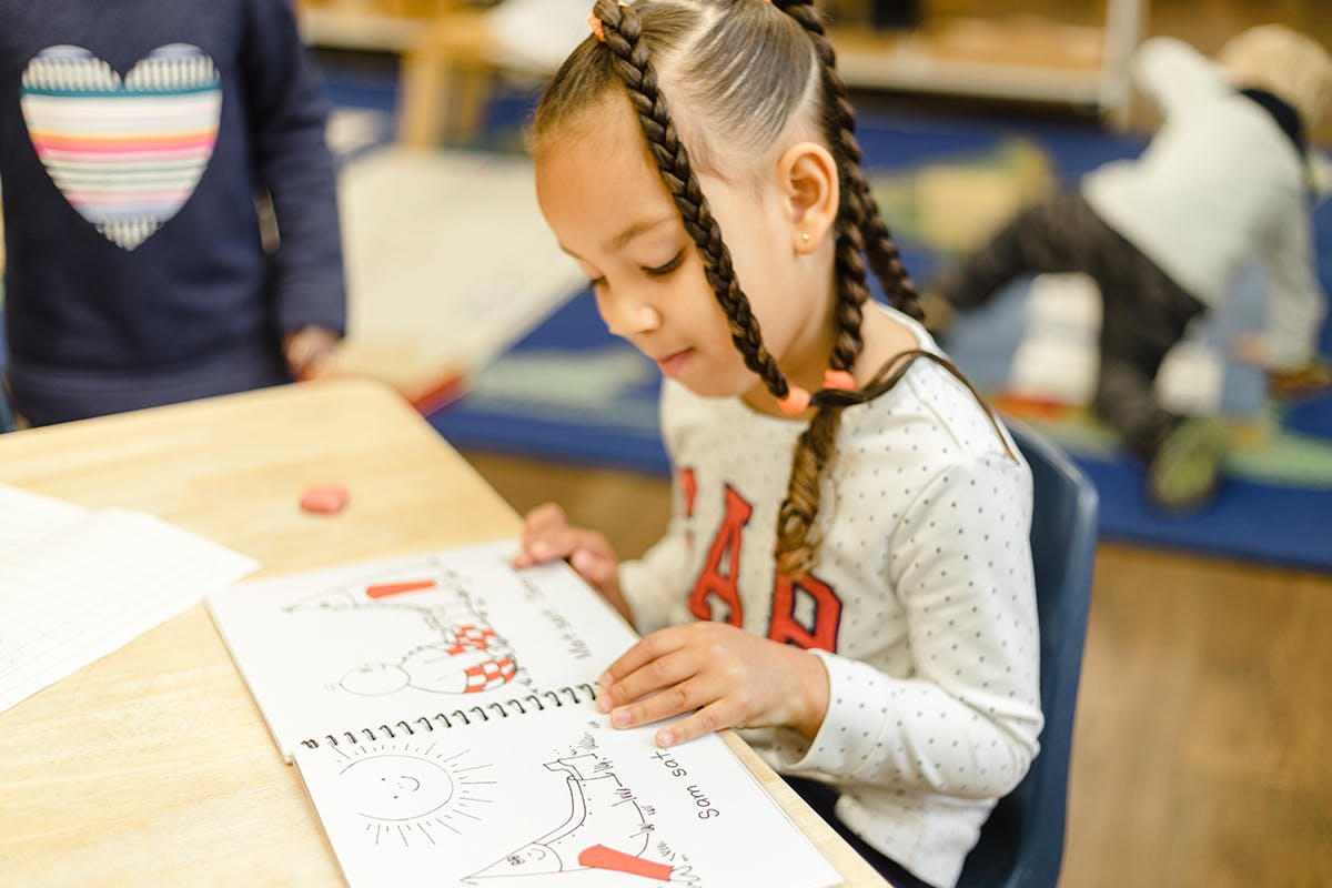 Child with braided hair looking at a book at a desk in a classroom