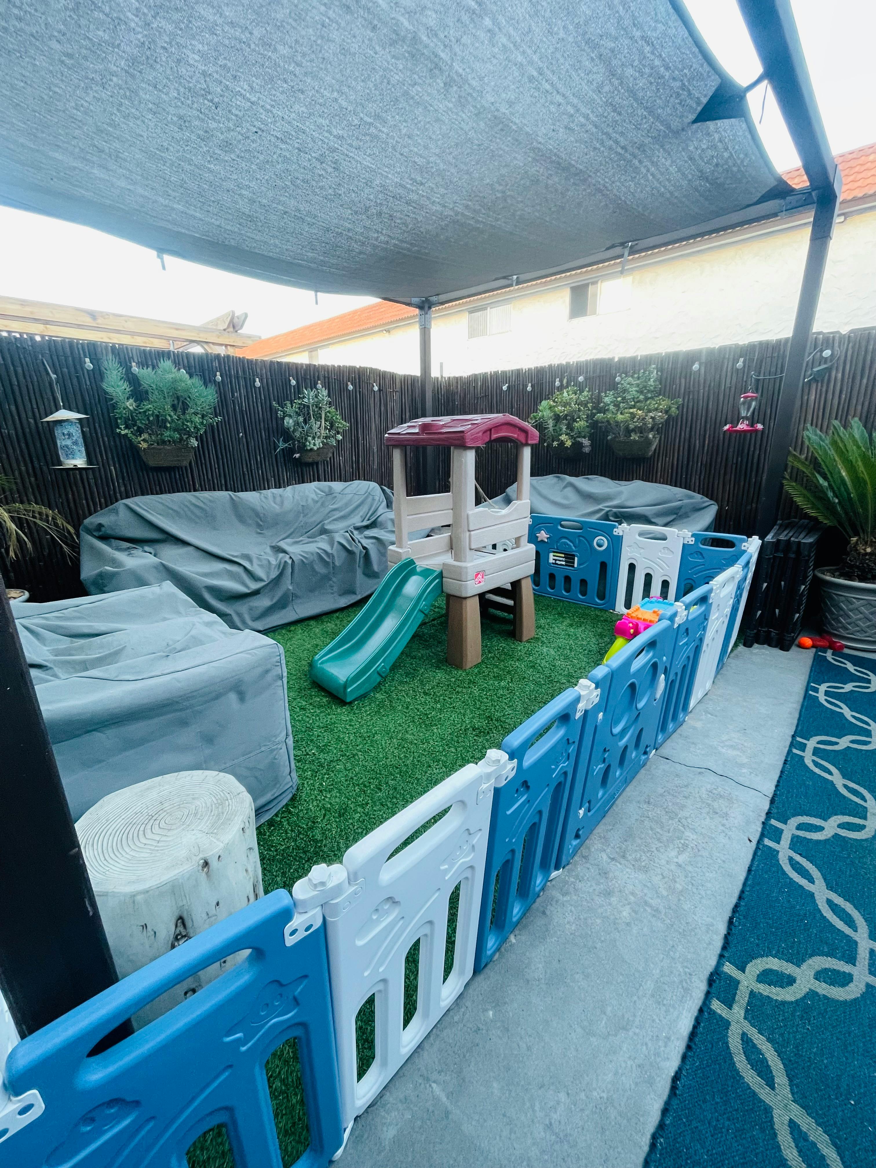 Outdoor play area with a small slide and play structure, fenced off from patio furniture