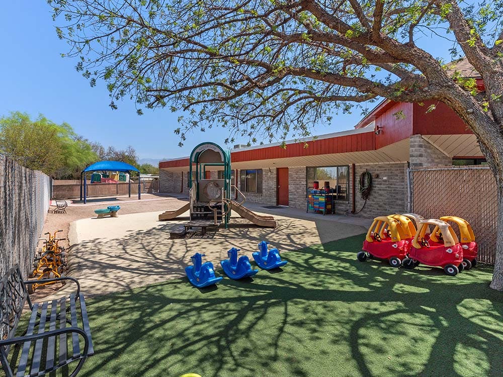 Outdoor playground with play structure, riding toys, and shade from a building