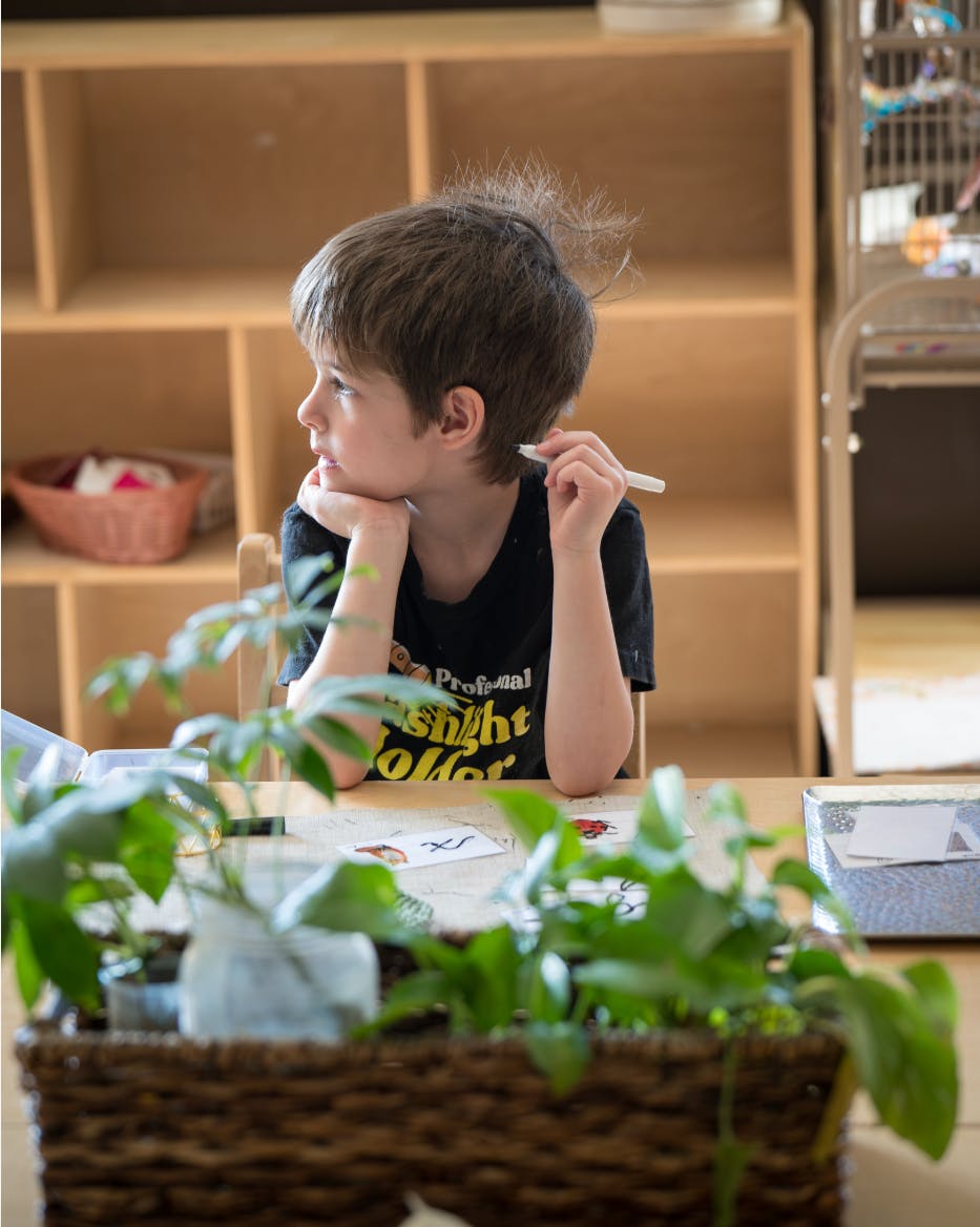 Child sitting at a table with chin resting on hand, holding a pen