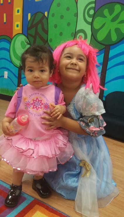 Two children, one in a pink dress holding a bottle, the other in a blue dress and pink wig