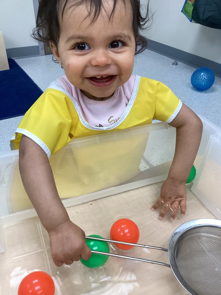 Child in a yellow bib playing with balls and a strainer in a container