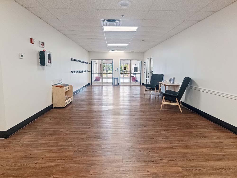 Empty hallway with wooden floor, benches on left wall, and two chairs on the right