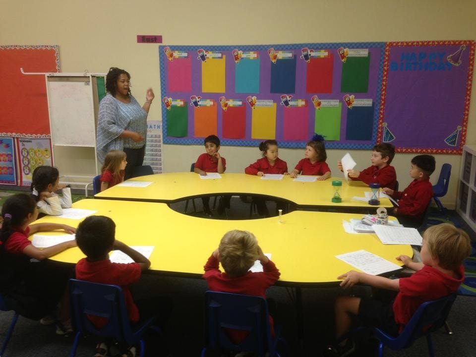 Adult speaking to children seated at a yellow table in a classroom