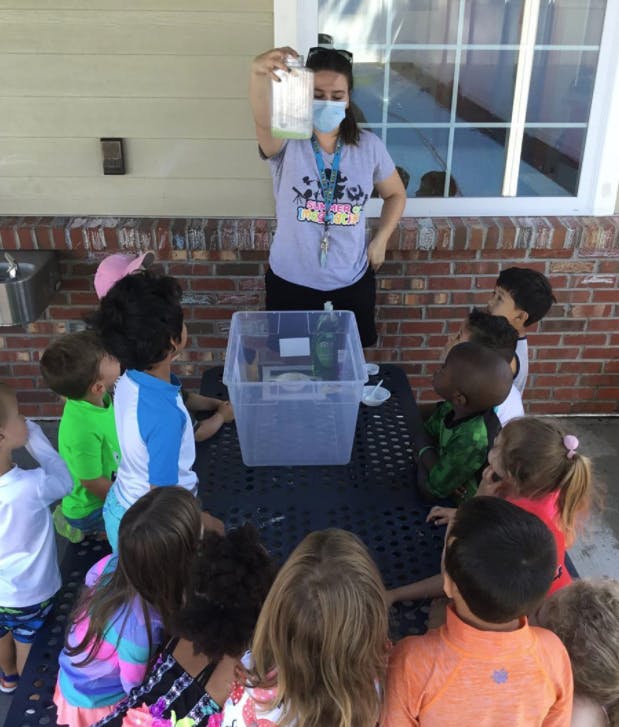 Person in mask holding up a jar for children gathered around a table