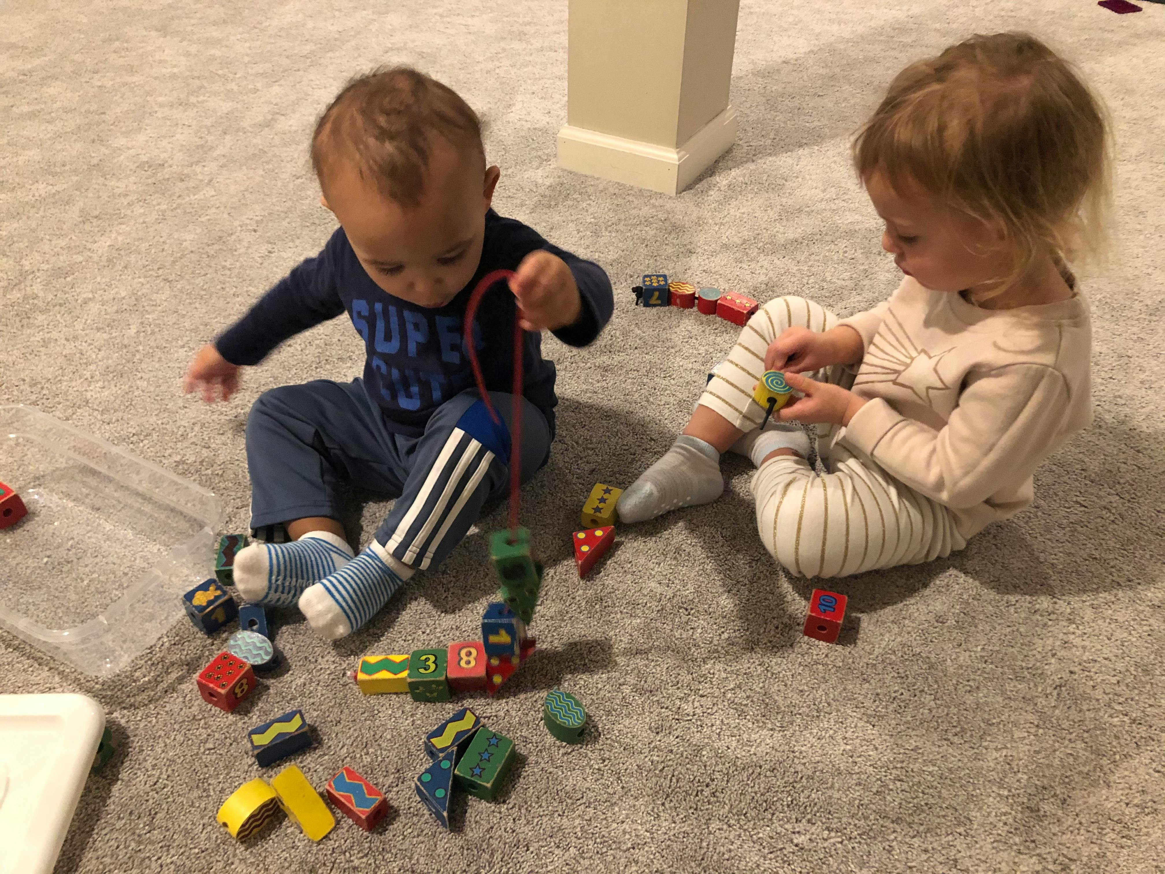 Two children playing with wooden blocks and a string on a carpeted floor