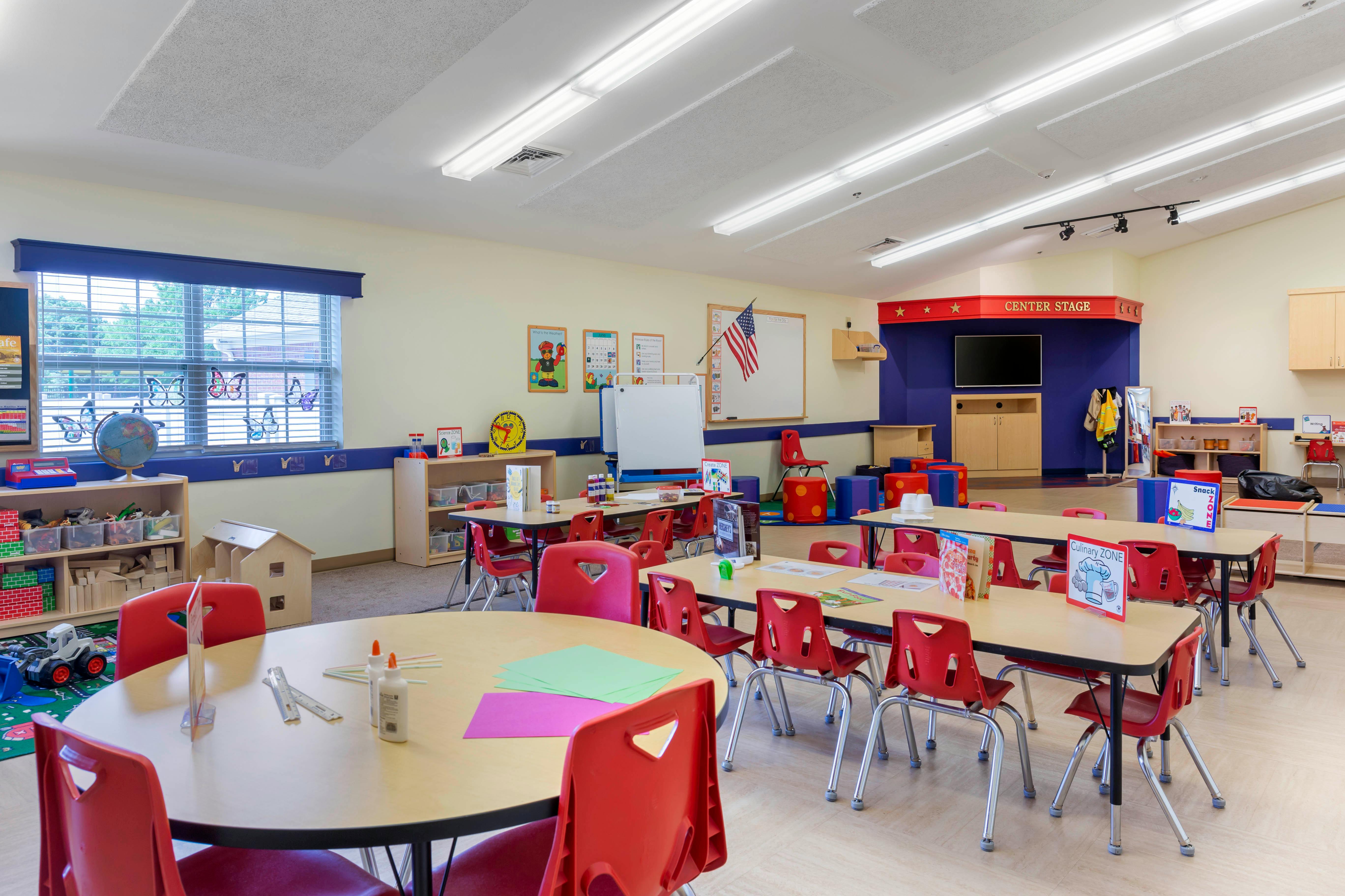 Classroom with small tables and red chairs, play areas, and a small stage