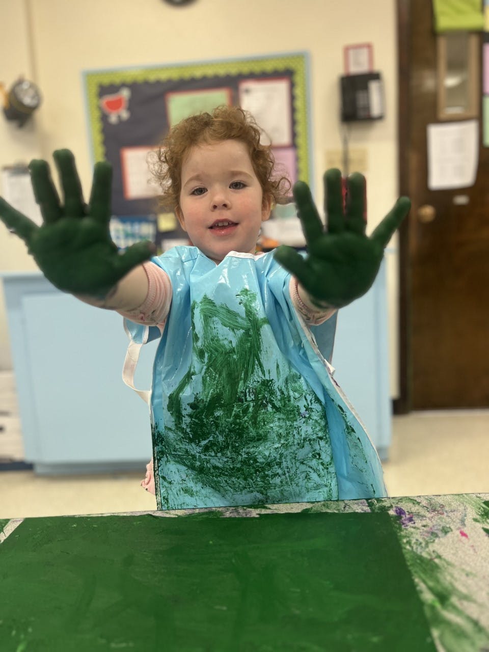 Child with green paint on hands and smock in a classroom