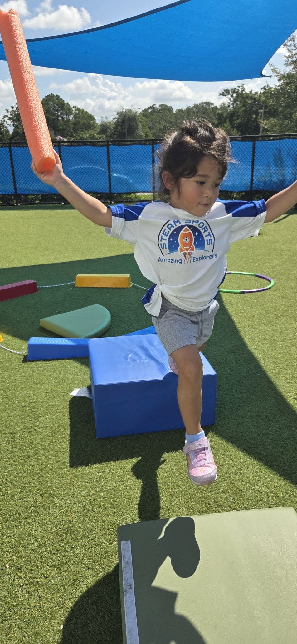 Child balancing on one leg on a blue block, holding an orange pool noodle