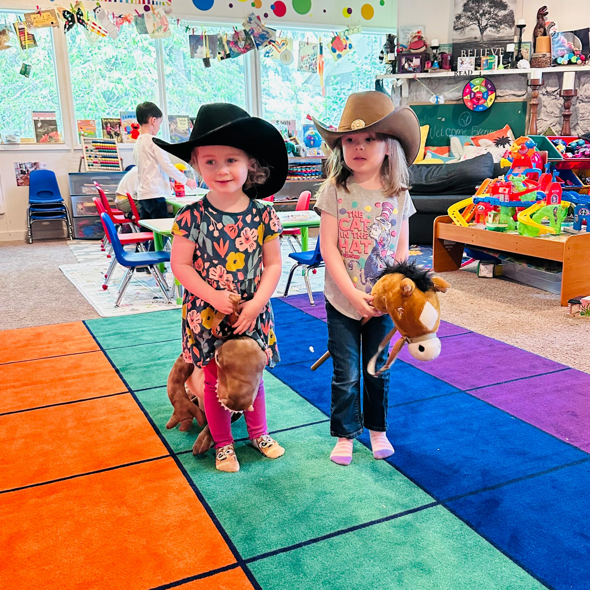 Two children wearing cowboy hats and holding toy horses in a classroom