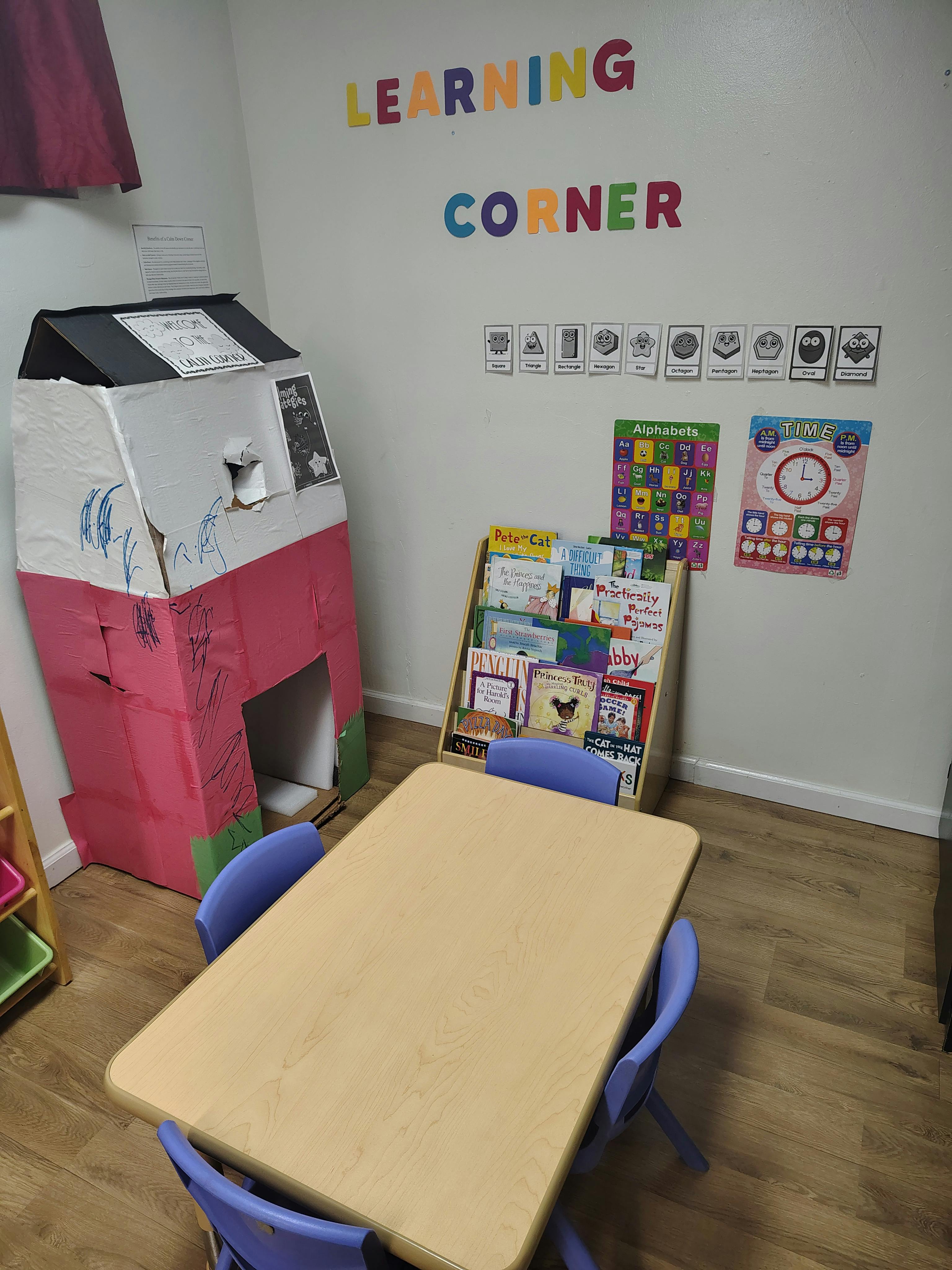 A classroom learning corner with a table, chairs, books, and a cardboard house