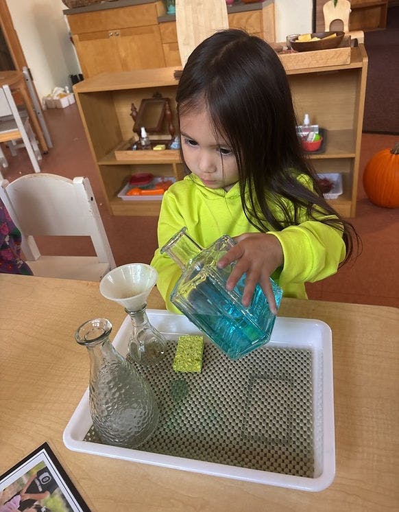 Child pouring blue liquid from a glass bottle into a funnel on a tray in a classroom