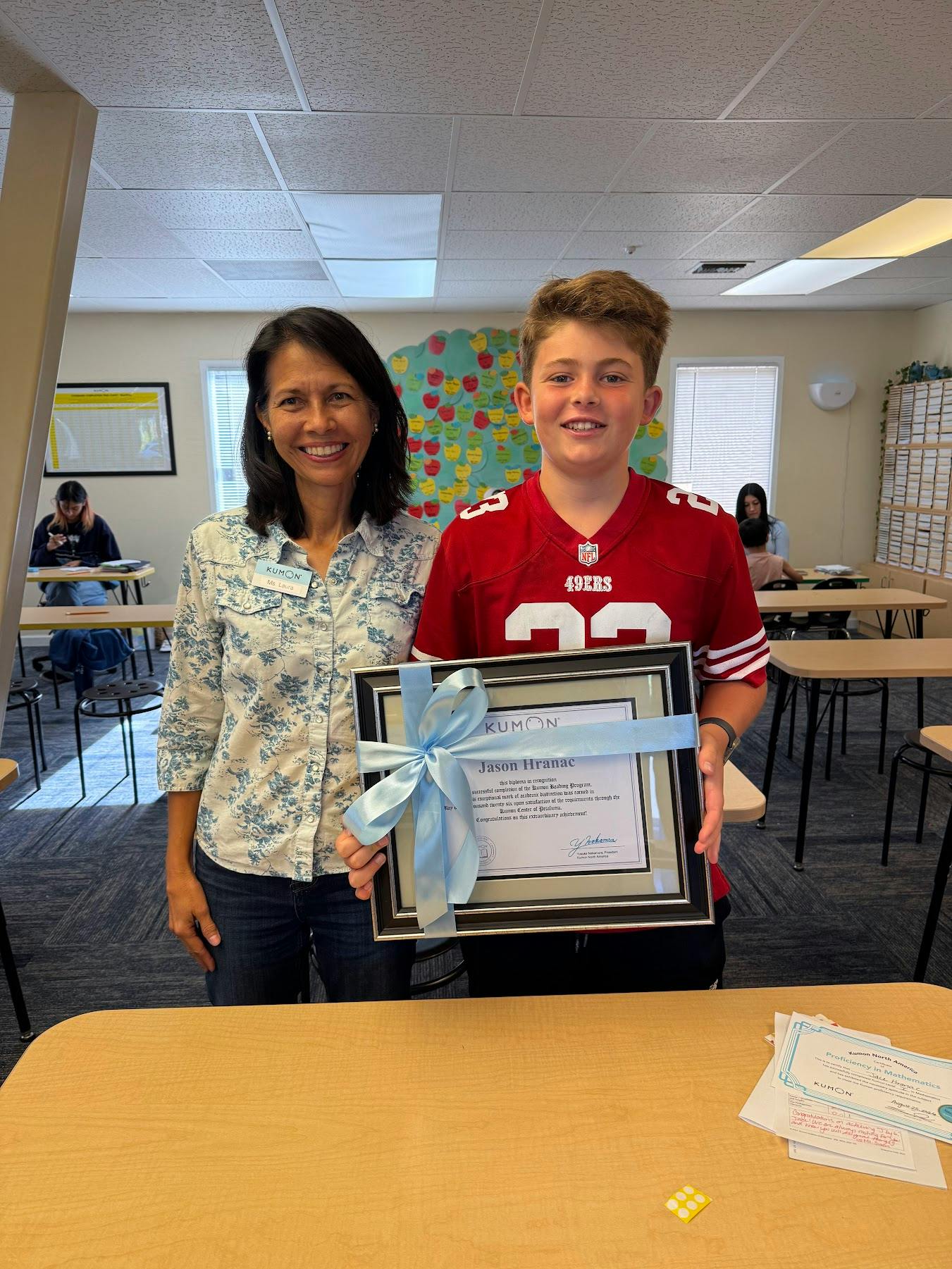 A person and a teen holding a framed Kumon certificate in a classroom