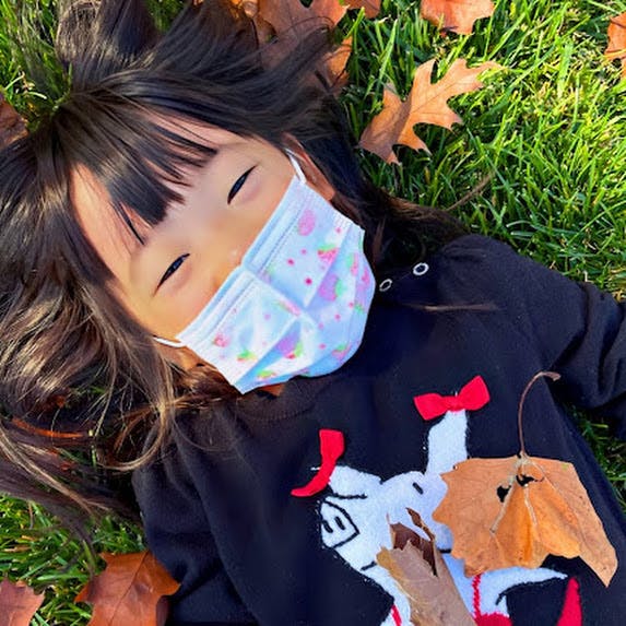 Child wearing a face mask lying on grass with autumn leaves