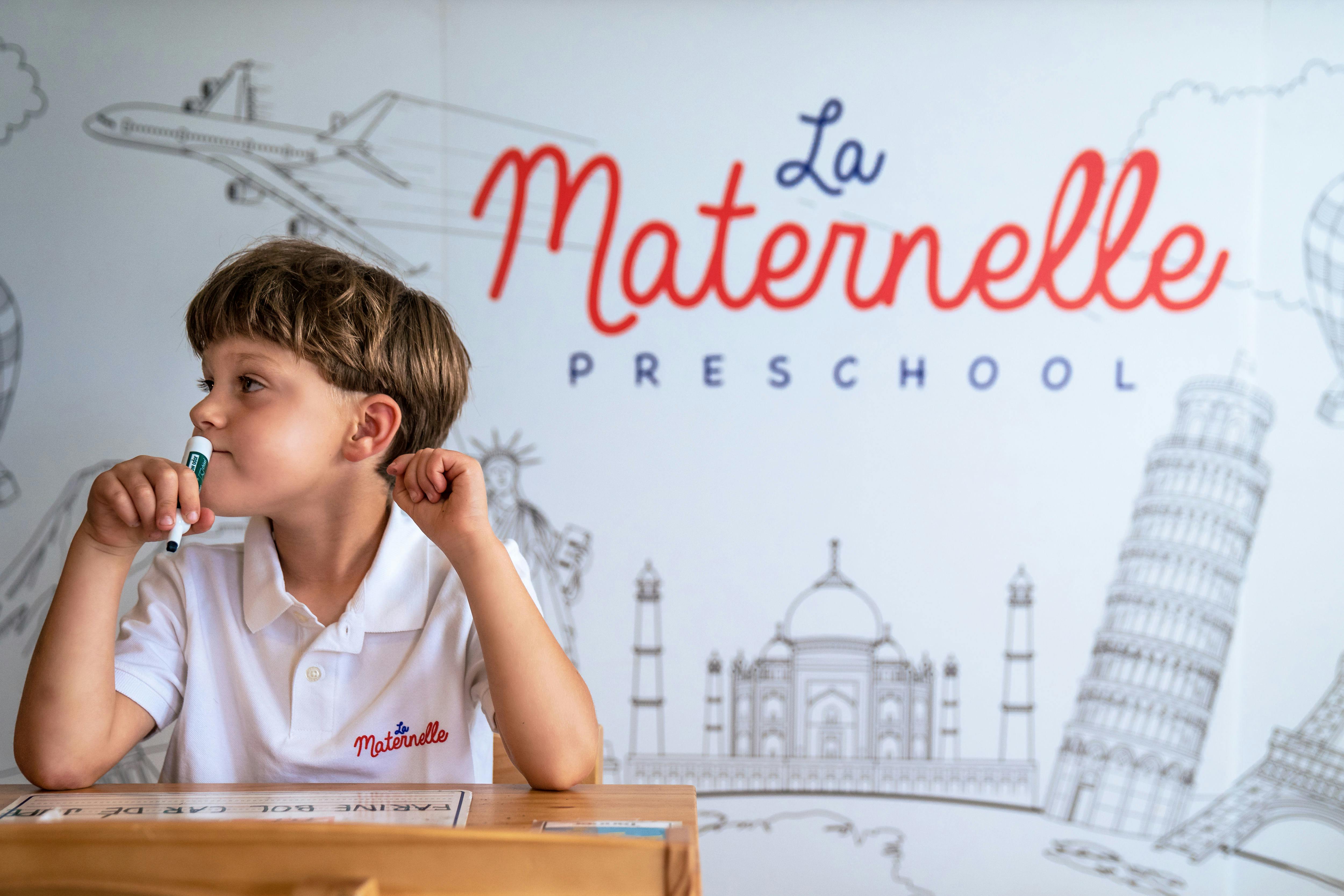 Child sitting at a desk holding a marker, with "La Maternelle Preschool" on the wall